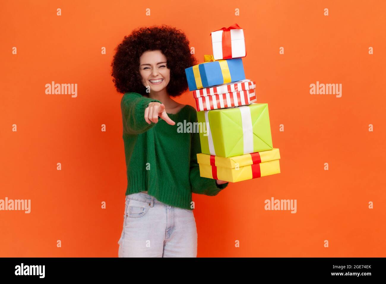 Beautiful woman with curly hair wearing green casual style sweater pointing finger to camera, holding stack of presents, choosing you as winner. Indoo Stock Photo
