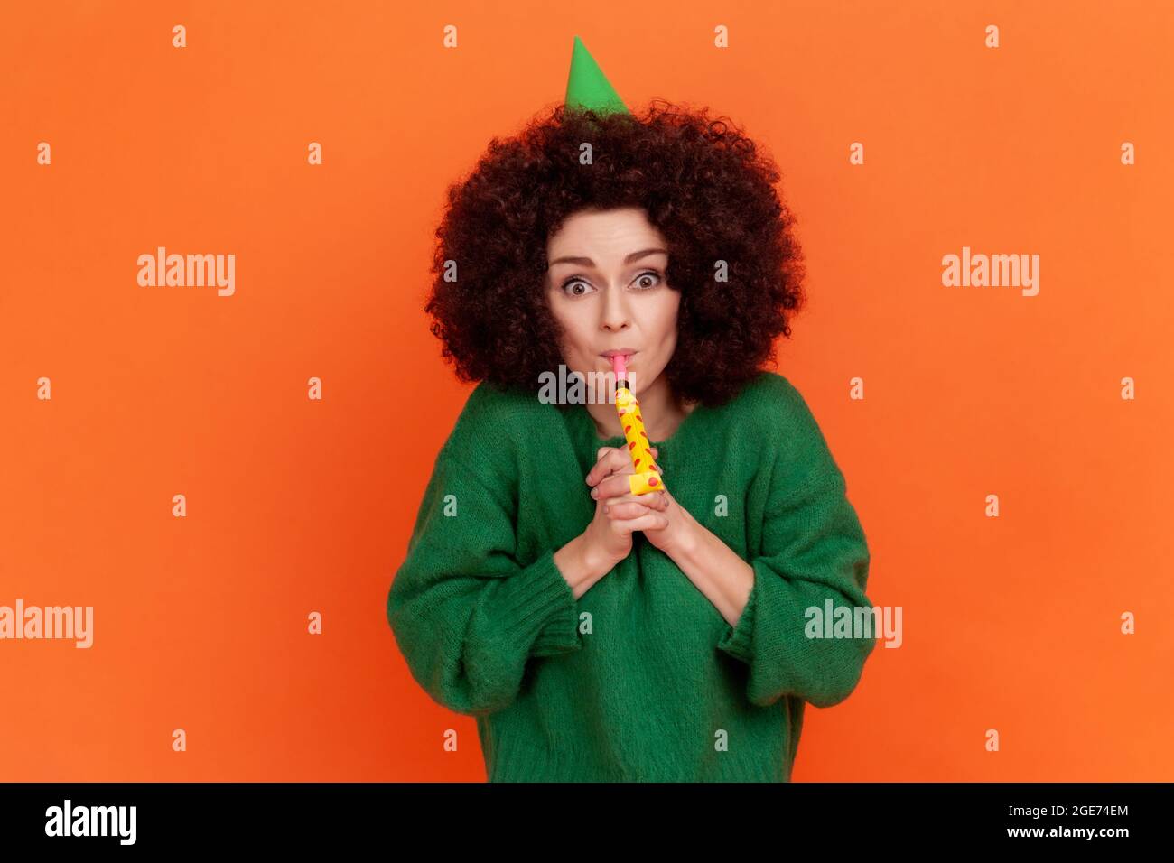 Happy birthday! Portrait of festive woman with curly hair wearing green casual style sweater and party cone blowing horn, celebrating holiday. Indoor Stock Photo