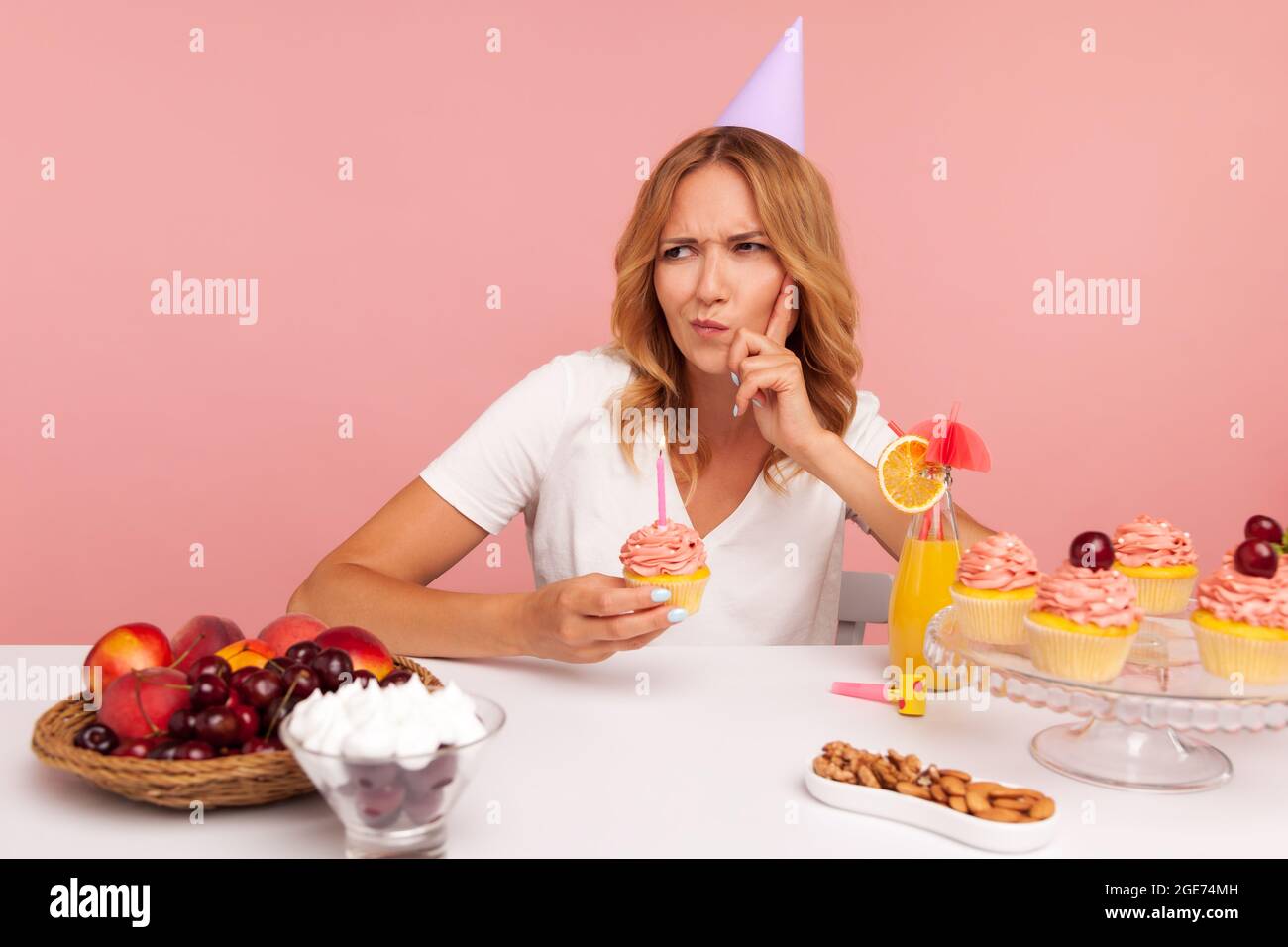 Thoughtful blonde woman wearing party cone thinking about birthday desire, making wish, frowning face and keeps finger in cheek, holds cake with candl Stock Photo