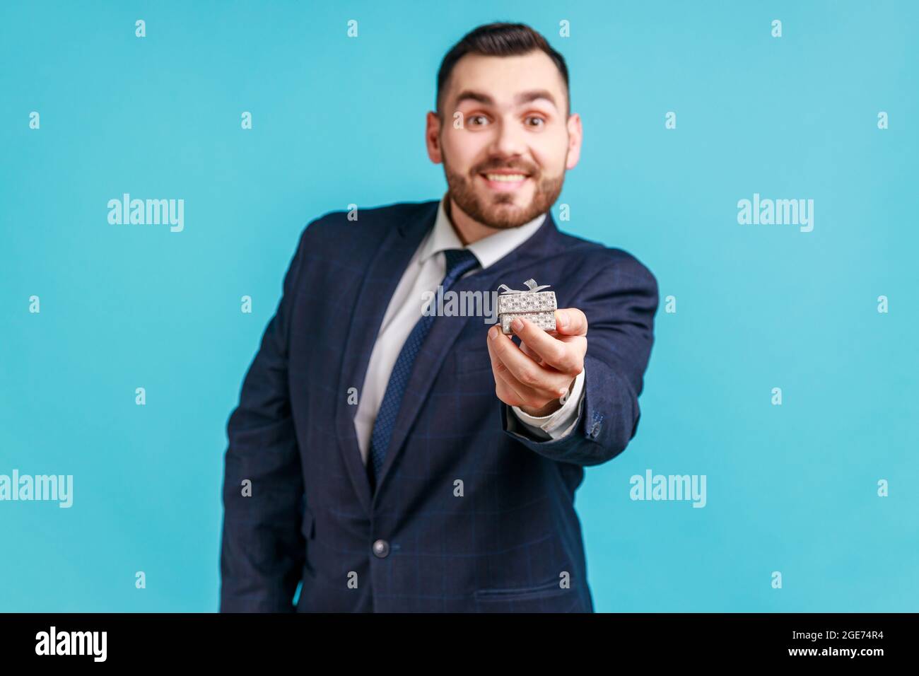 Happy smiling man wearing dark official style suit holding out little box with ring, making proposal to her girlfriend, looking at camera. Indoor stud Stock Photo