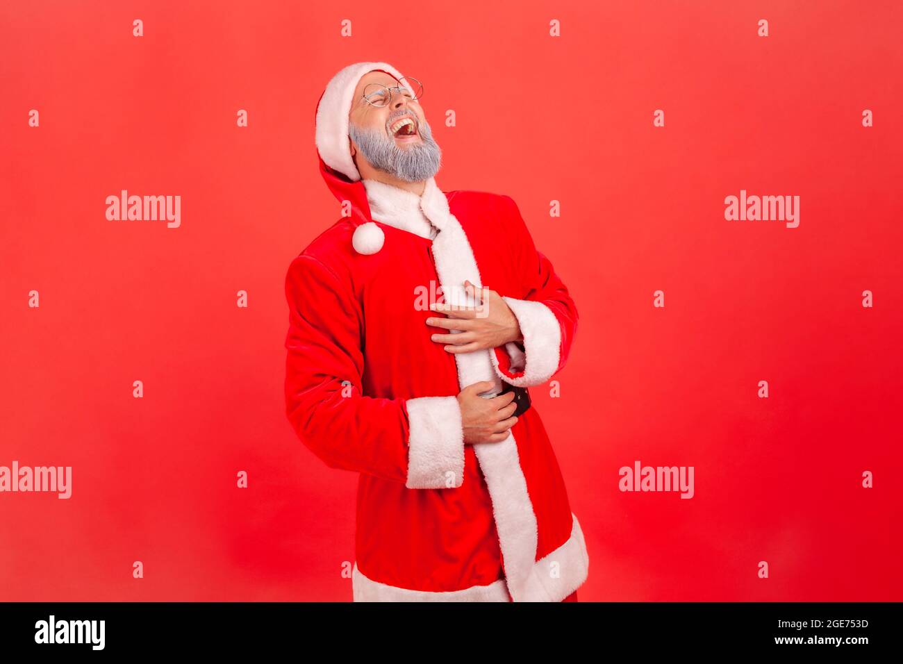 Portrait of elderly man with gray beard wearing santa claus costume holding hands on belly and can't stop laughing hard, hearing funny stories, joke. Stock Photo