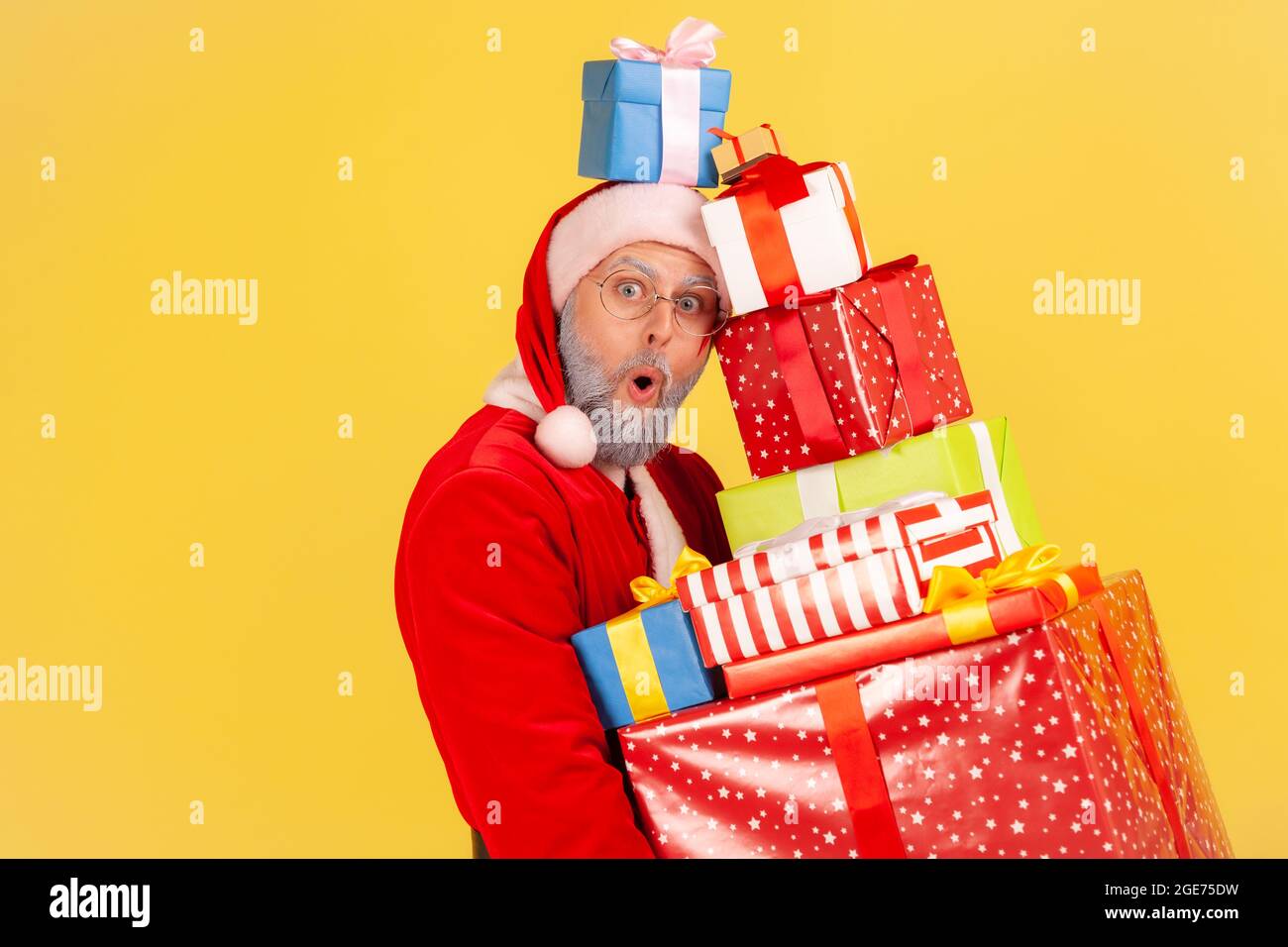 Shocked elderly man with beard in santa claus costume standing with many Christmas gifts, looking at camera with astonished face, heavy presents box. Stock Photo
