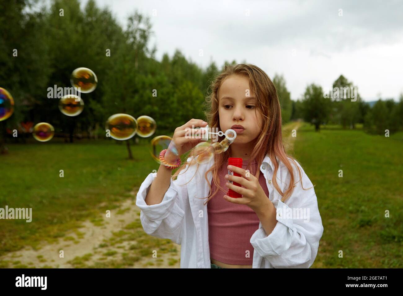 Cute girl in casual clothes blowing soap bubbles while standing on path on overcast summer day in nature Stock Photo