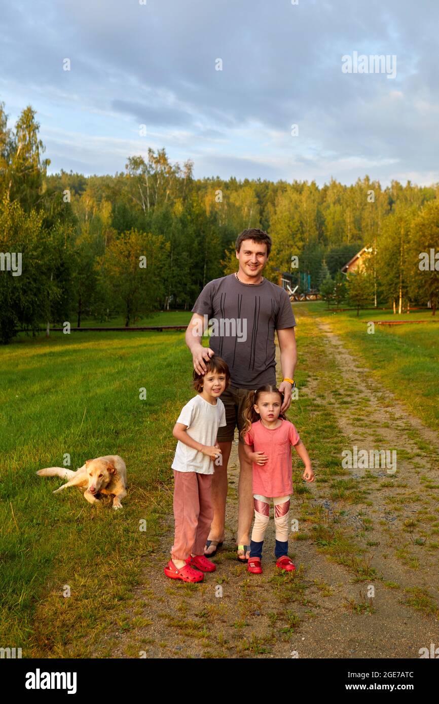 Adult man with daughter and son standing on path near loyal dog on summer weekend day in nature Stock Photo