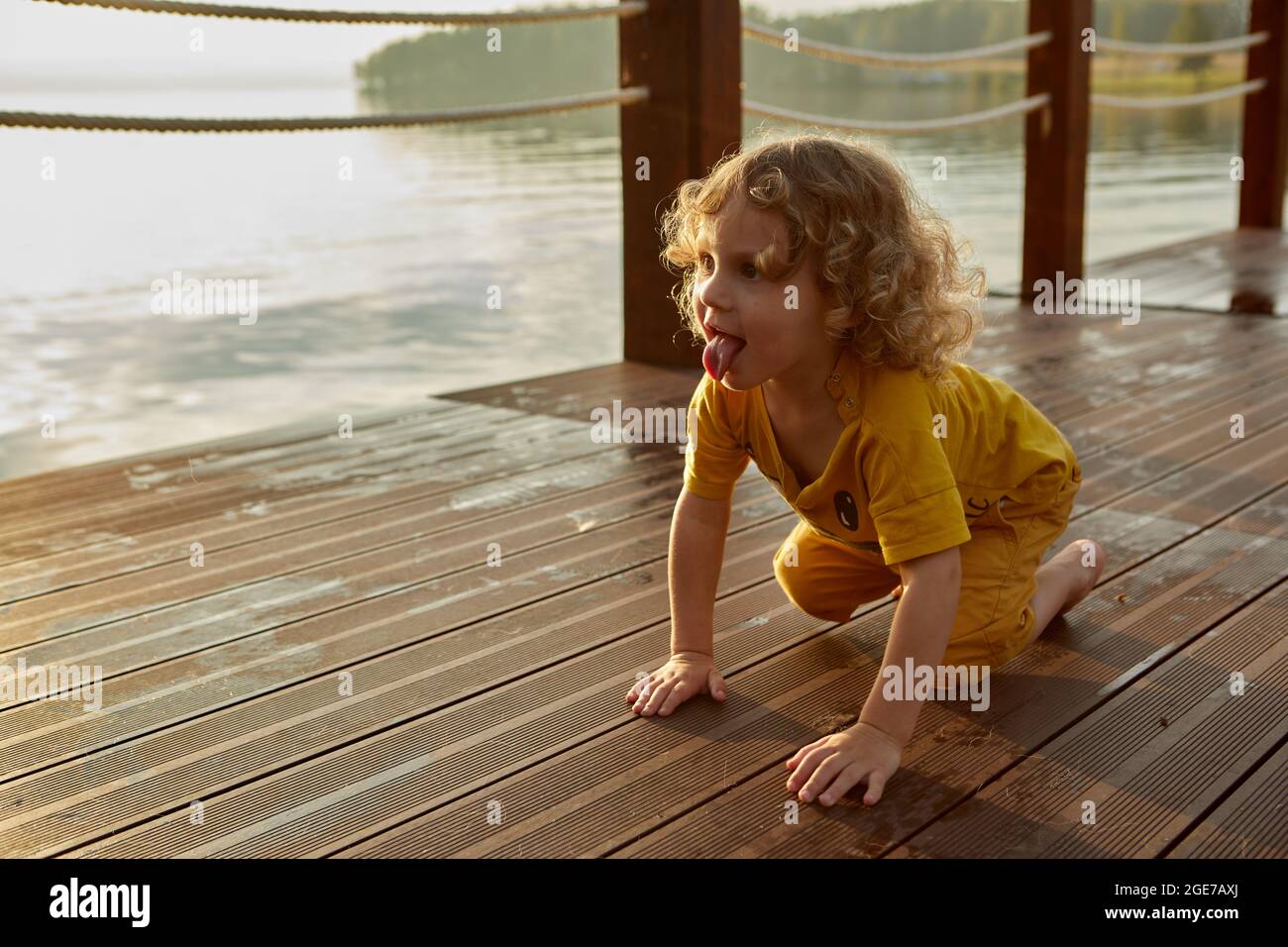 Adorable toddler with curly hair showing tongue and crawling on wet quay near lake water at sunset Stock Photo