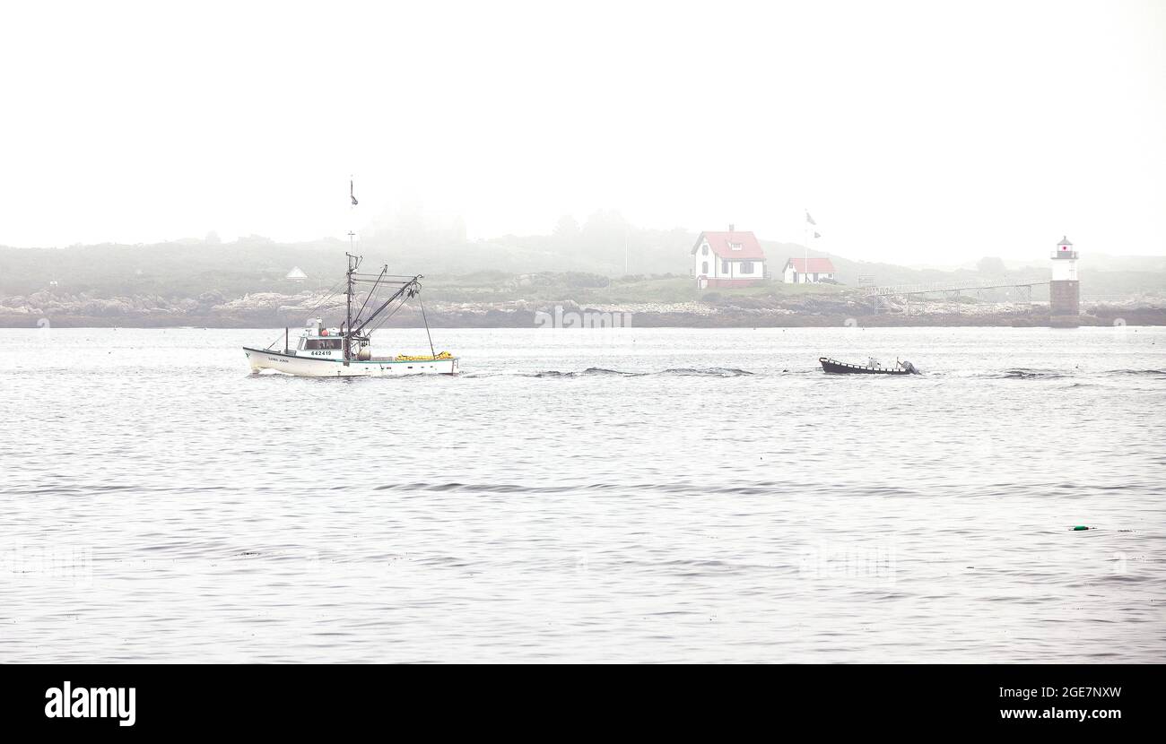 A commercial fishing boat with a dory under tow passing Ram Island Light, off Ocean Point (Boothbay),Maine, USA, in a early morning fog Stock Photo