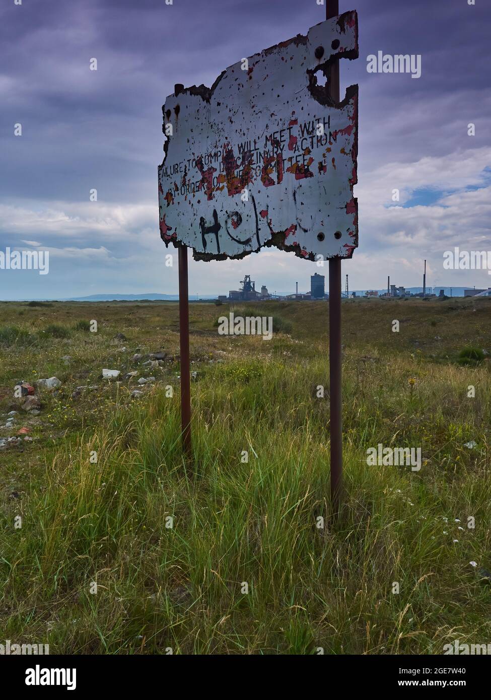 An illegible, disintegrating sign in the slag fields, its posts framing the abandoned Redcar steel mill on the horizon, under a dramatic clouded sky. Stock Photo