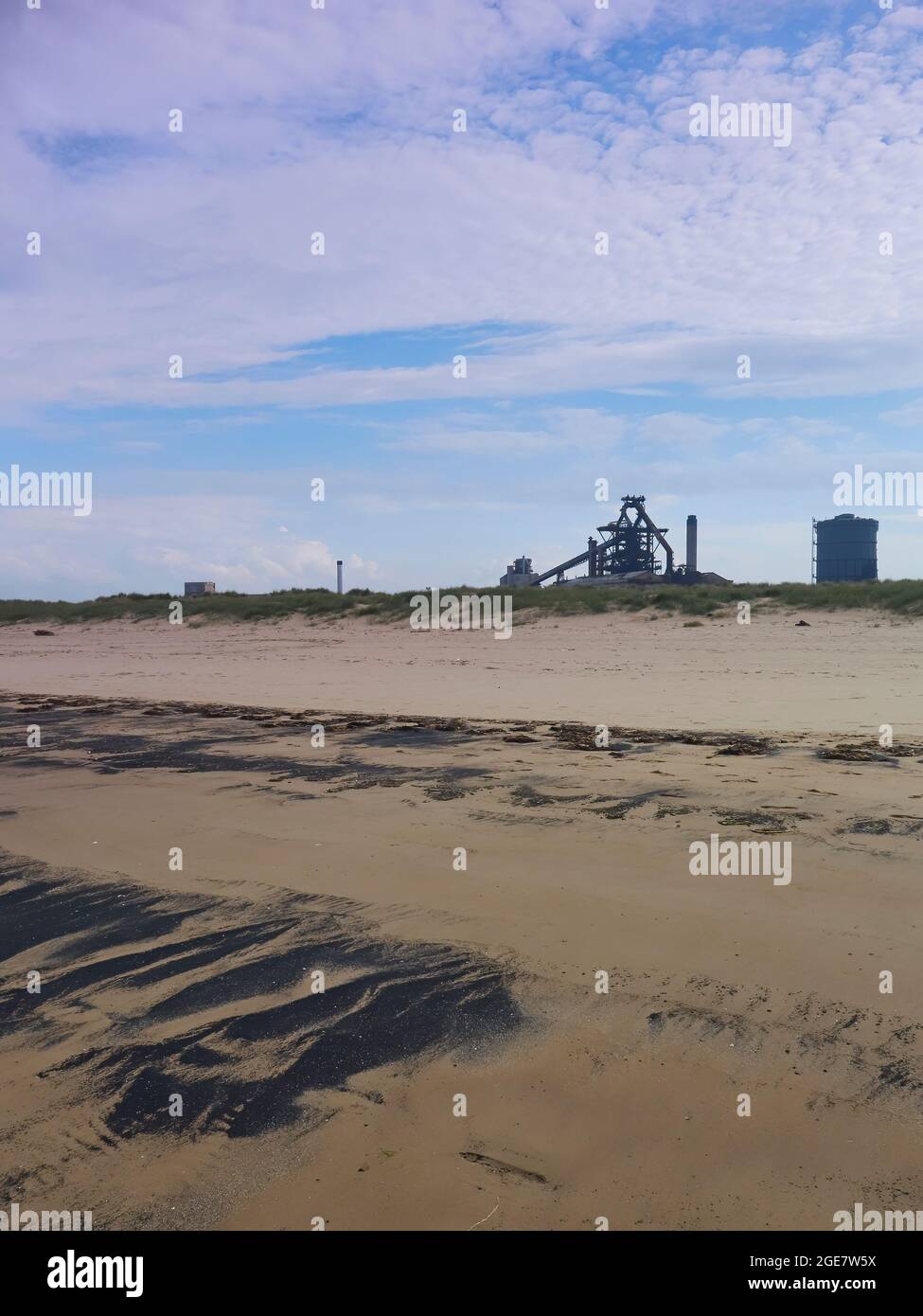 A sweep of sea coal, sand and sand dunes, ahead of the skeletal silhouette of Redcar’s abandoned steel works, under fluffy clouds and blue summer sky. Stock Photo
