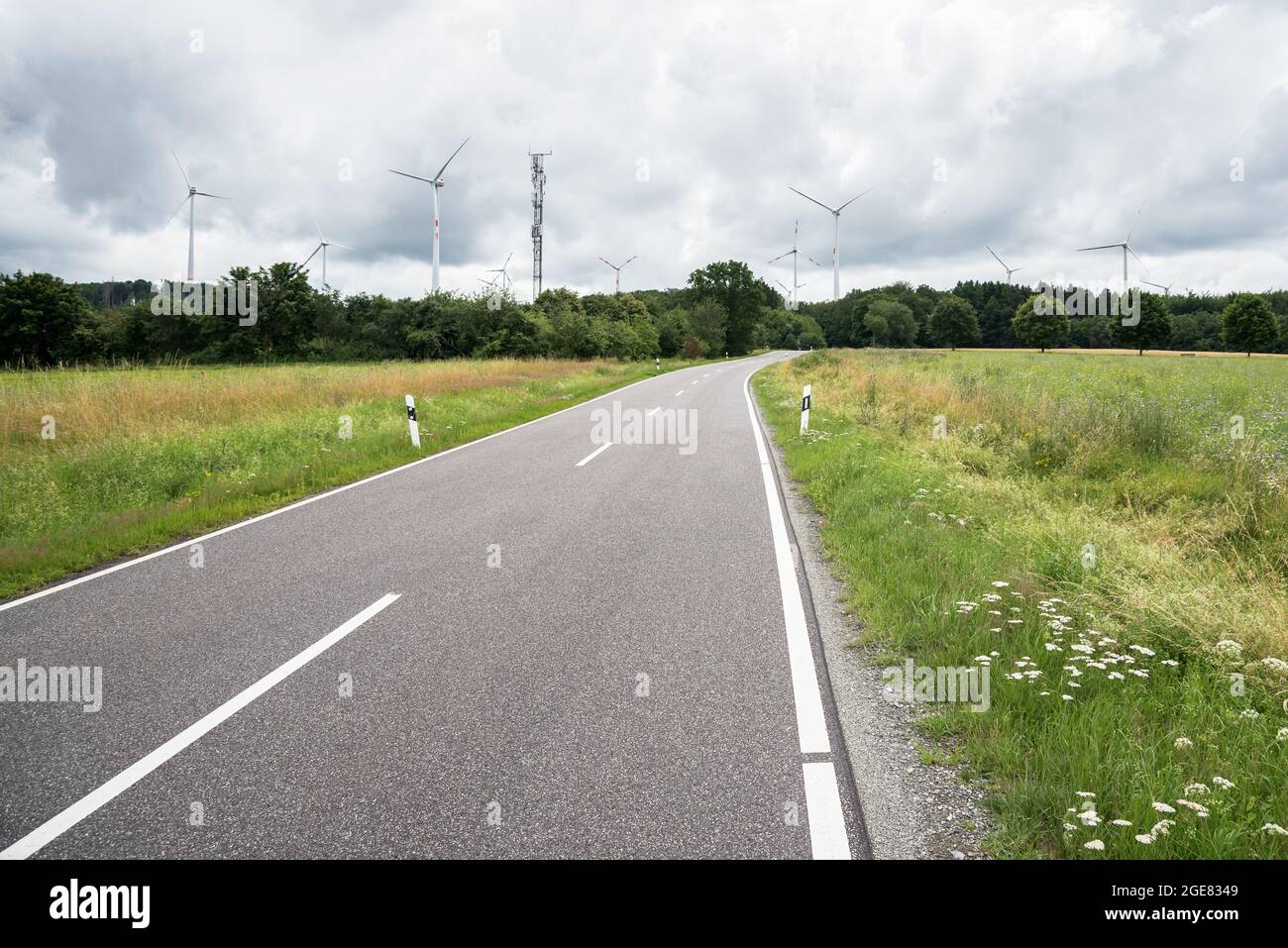 Deserted road through a meadow with wind turbines in background on a cloudy summer day Stock Photo