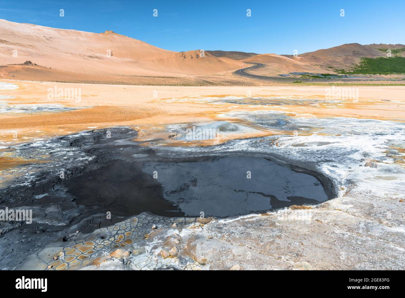 Boiling mud pool in a geothermal area in Iceland on a sunny summer day. A winding road  climbing a barren volcanic hill is visible in background. Stock Photo