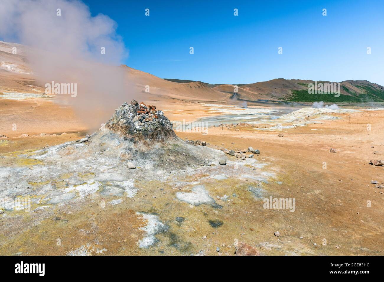 Fumarole in a geothermal area on a clear summer day. A winding road up a barren hill is visible in background. Stock Photo