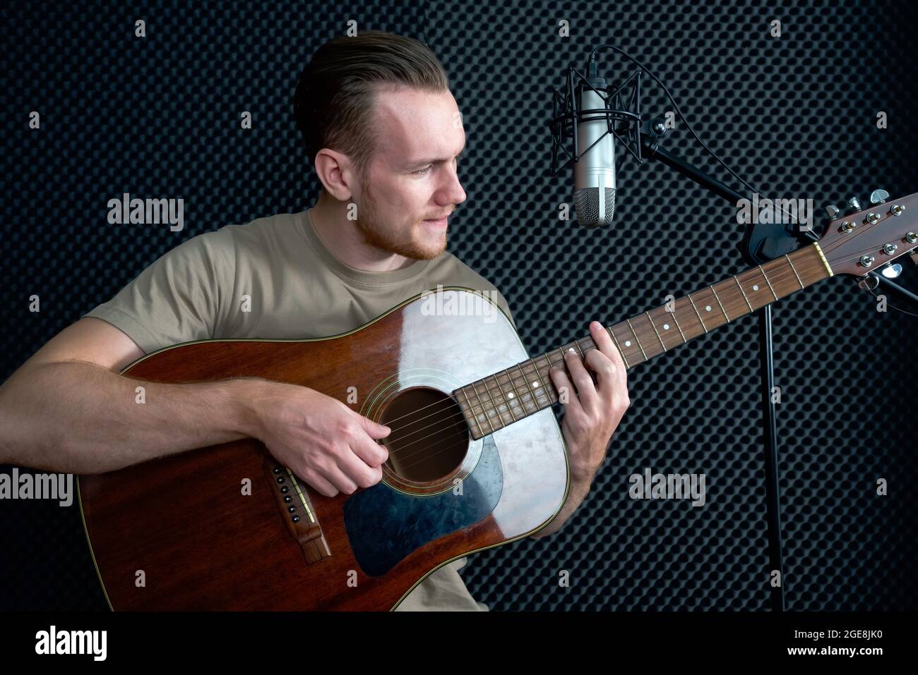 Young caucasian man sings while playing an acoustic guitar in front of black soundproofing walls. Musicians producing music in professional recording Stock Photo