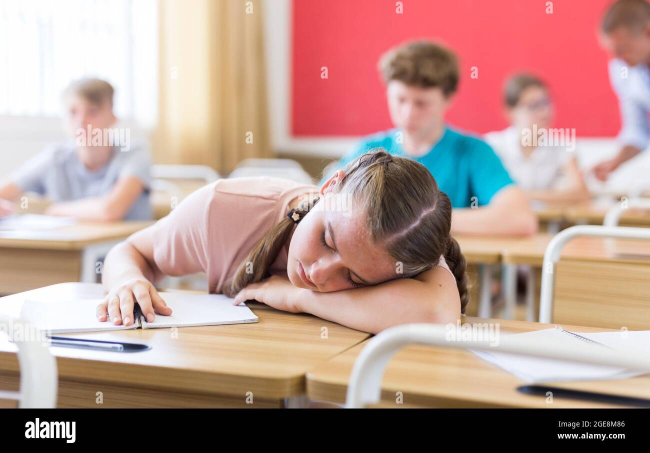 Tired teenage girl sleeping at desk in classroom during lesson Stock