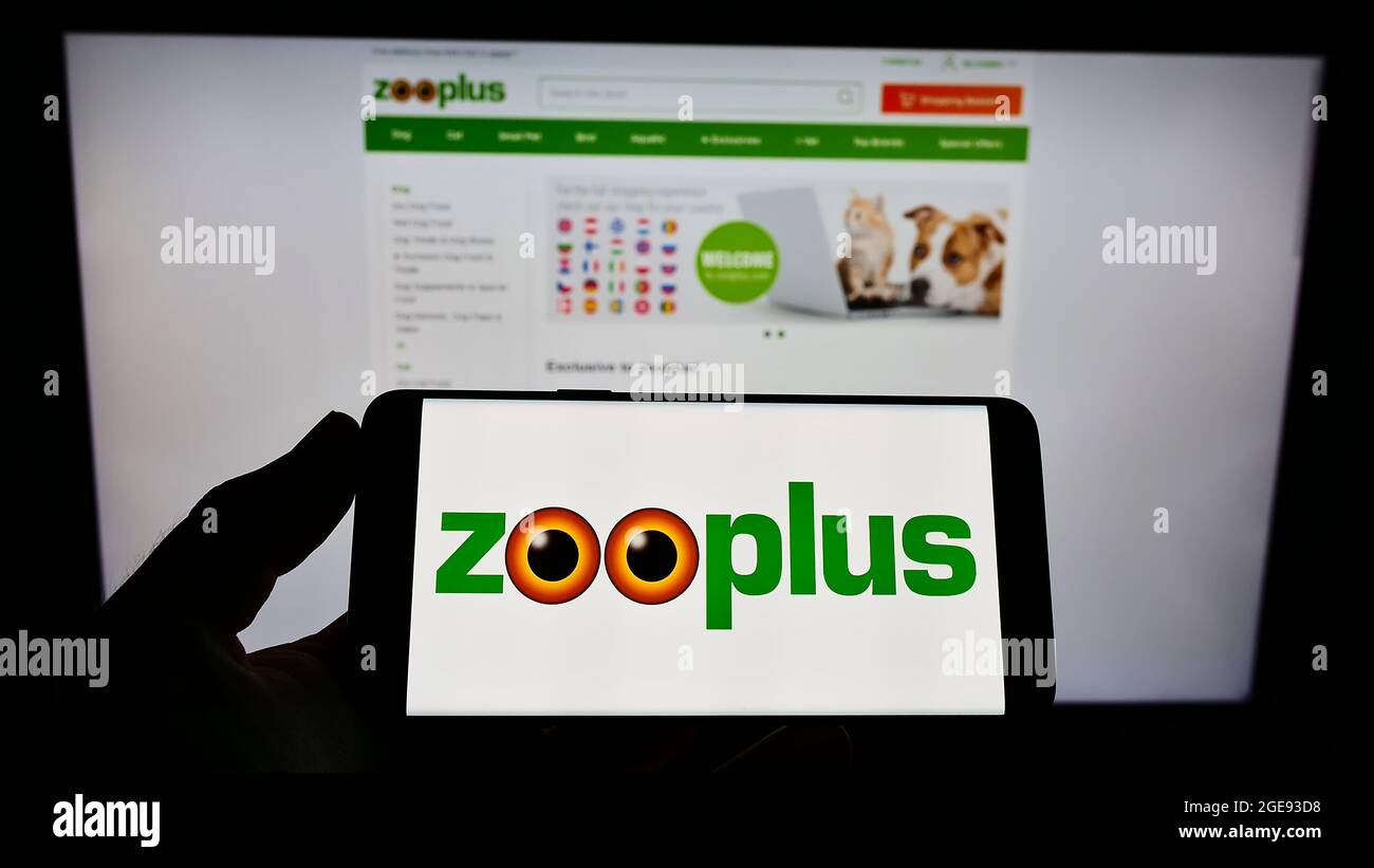 Person holding smartphone with logo of German pet food retail company Zooplus AG on screen in front of website. Focus on phone display. Stock Photo