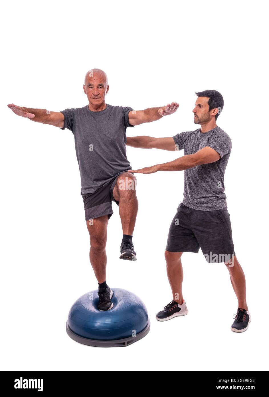 An elderly man doing a balance exercise, on one leg, on a hemisphere ball. With help of a fitness trainer. On a white isolated background. Stock Photo