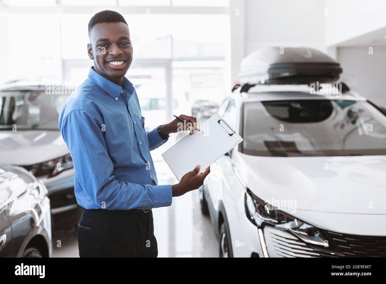 Cheerful black car salesman holding empty clipboard, smiling at camera in modern auto dealership, mockup Stock Photo