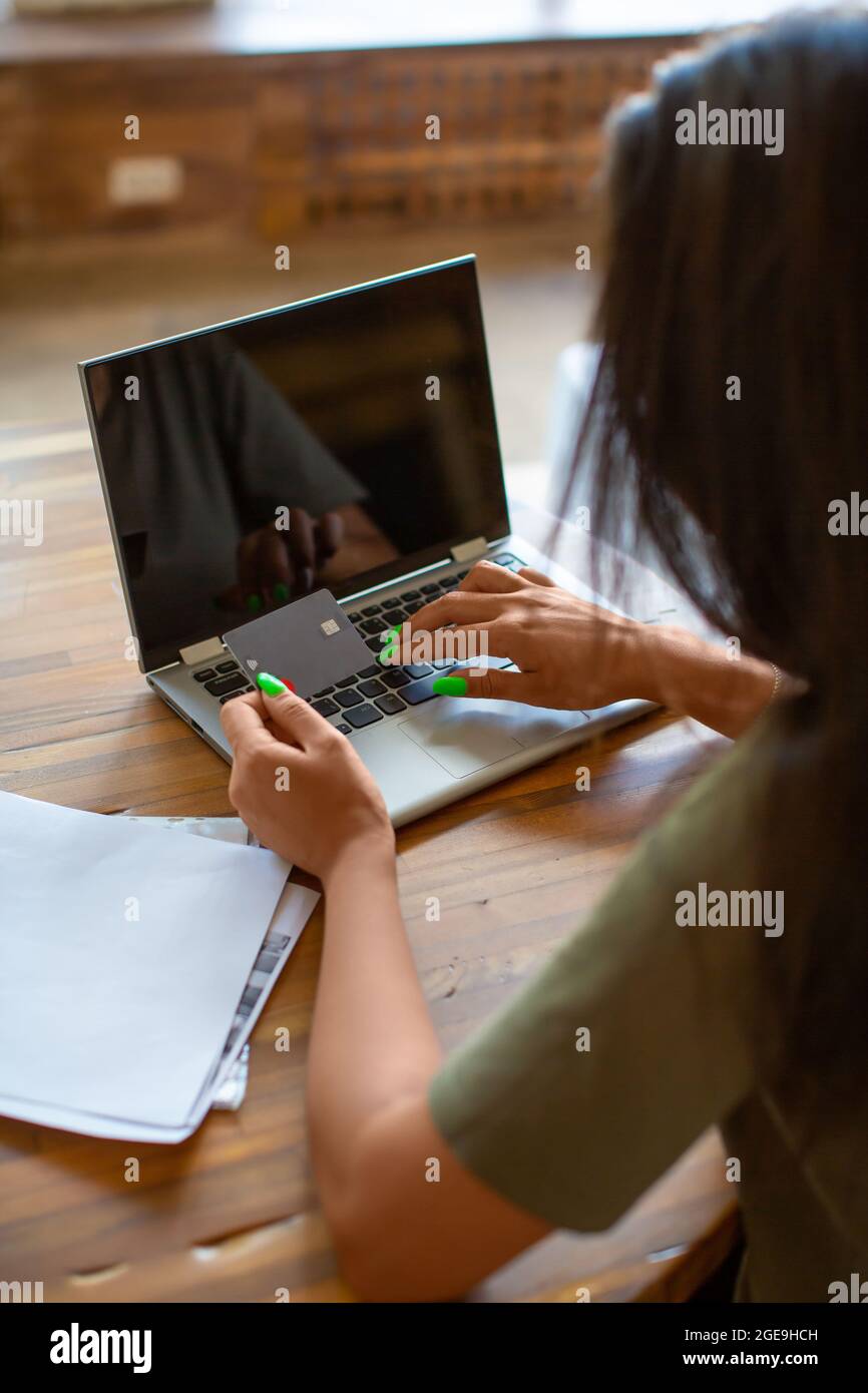 Back view of a woman with credit card in hands in front of laptop monitor. Online shopping at home concept. Vertical photo. Stock Photo