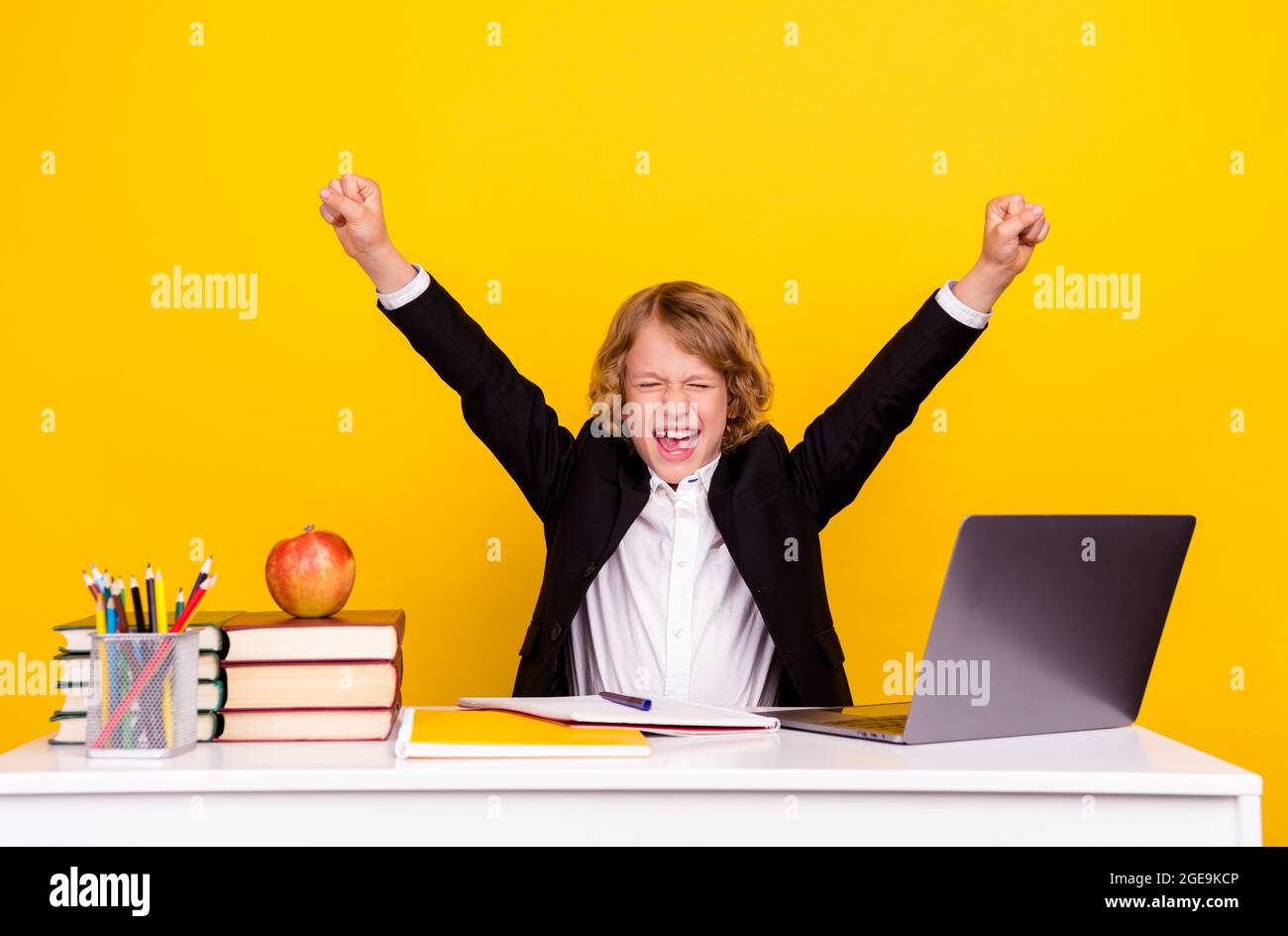 Photo of astonished child sit desk scream celebrate victory raise fist wear uniform isolated yellow color background Stock Photo