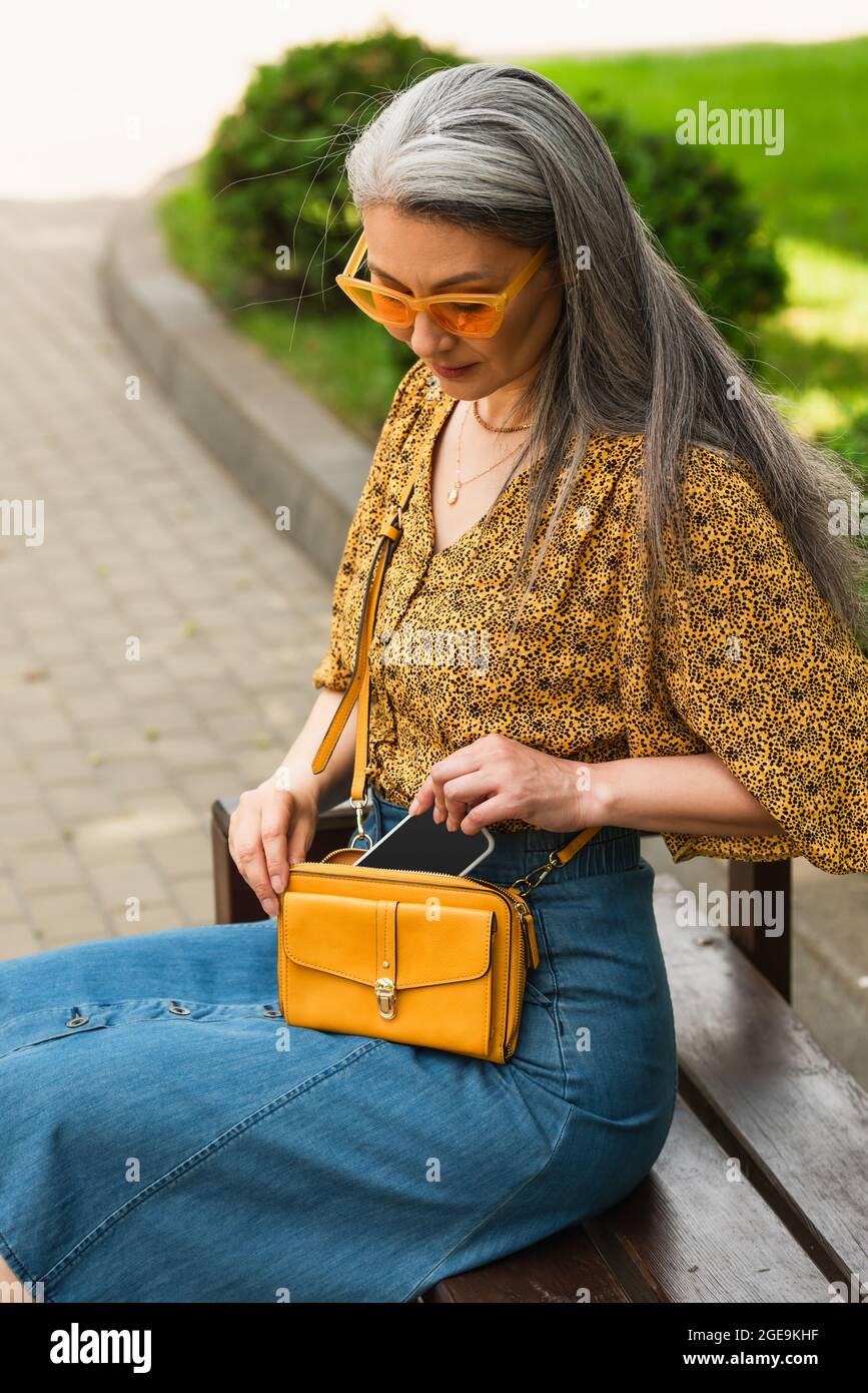 trendy mature woman with crossbody bag and smartphone sitting on