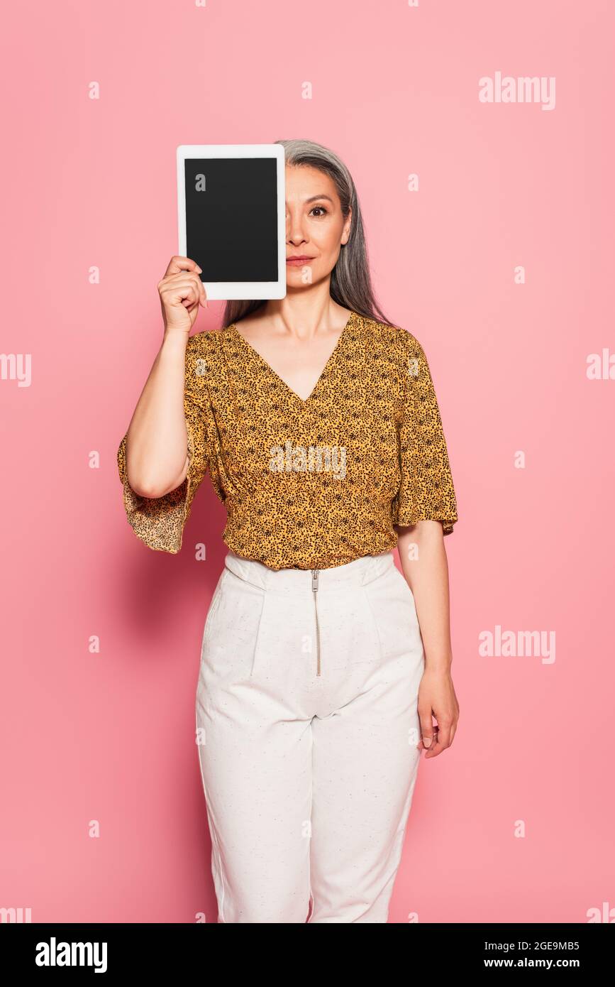 middle aged woman in patterned blouse covering face with digital tablet on pink background Stock Photo
