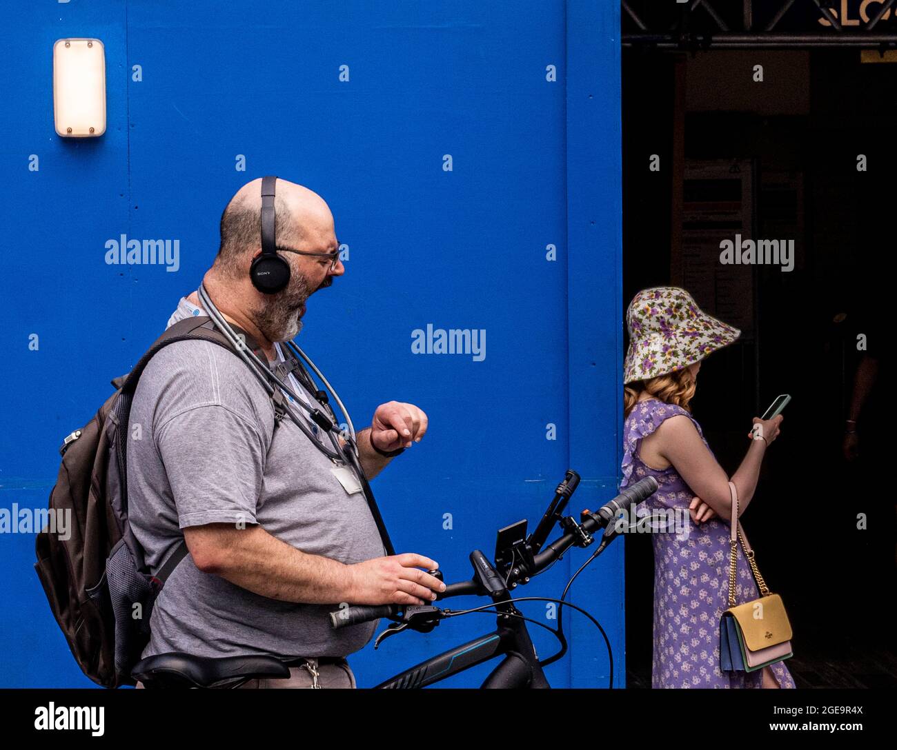 A man wearing a bike lock around his neck yawns really widely against the background of a blue door with a woman leaning on it in Sloane Square in Central London. Stock Photo