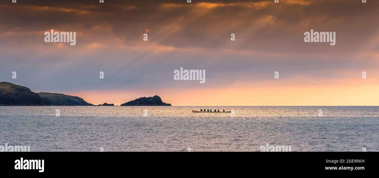 A panoramic image of a traditional historic Cornish Pilot Gig being rowed as the sun sets over Fistral bay in Newquay in Cornwall. Stock Photo