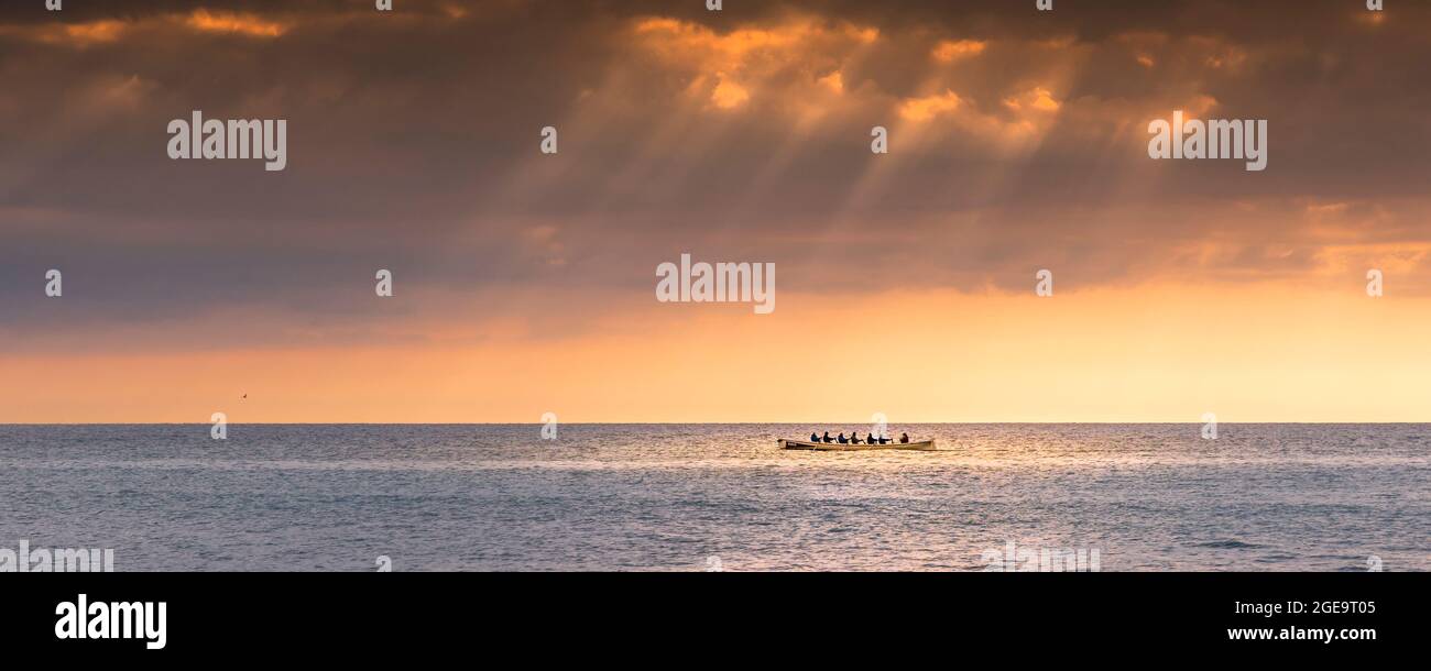 A panoramic image of the 200 year old Cornish Pilot Gig Dove being rowed as the sun sets over Fistral bay in Newquay in Cornwall. Stock Photo