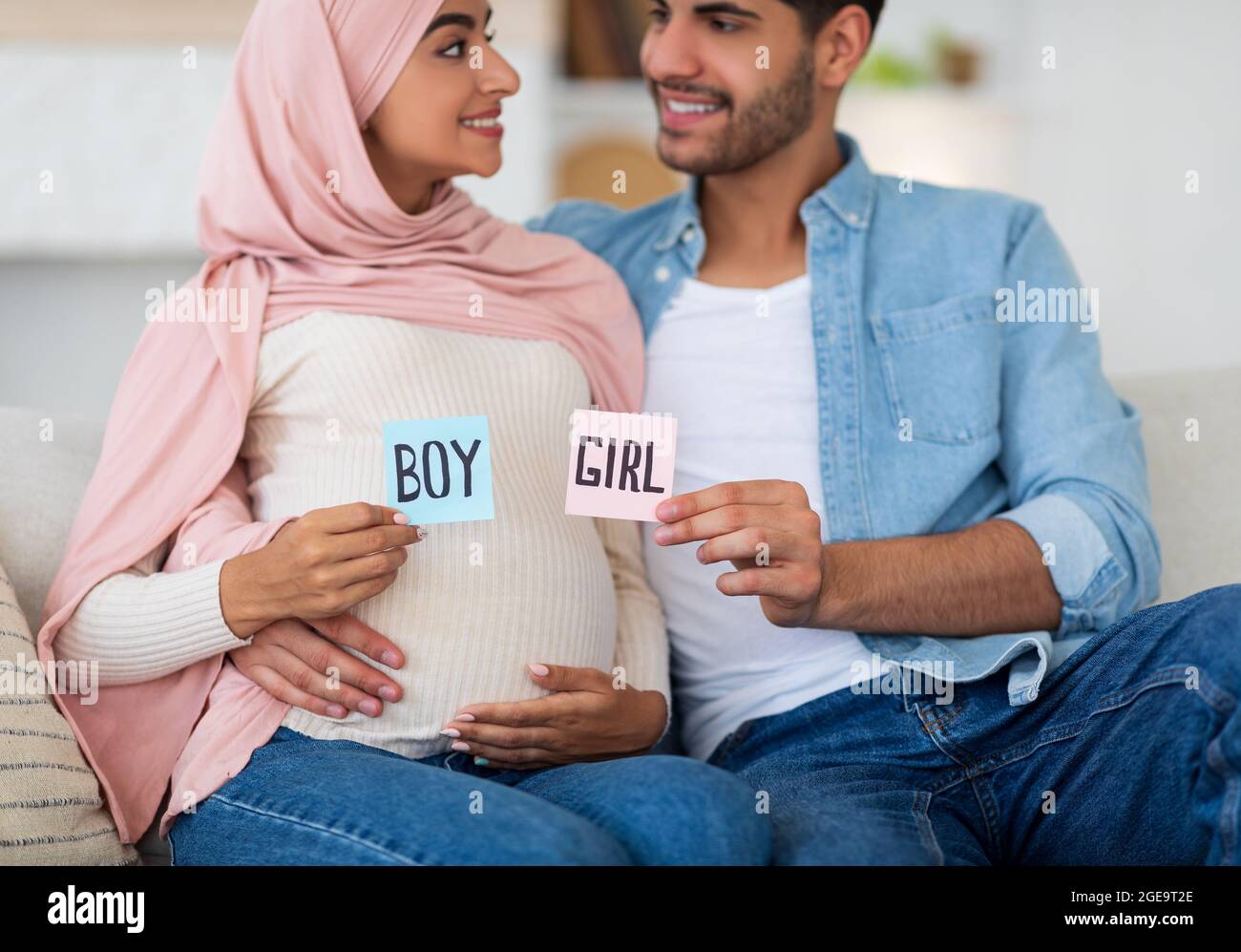 Gender reveal concept. Happy arab couple holding boy and girl sticker cards in pink and blue colors, sitting on couch Stock Photo