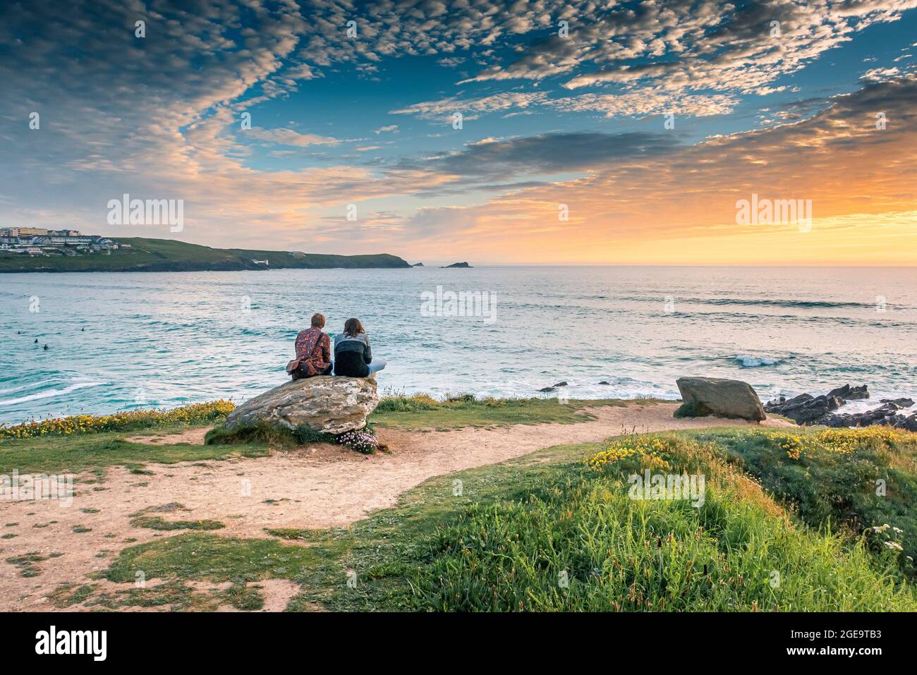 A young couple sitting on a rock overlooking Fistral as the sun sets in Newquay in Cornwall. Stock Photo