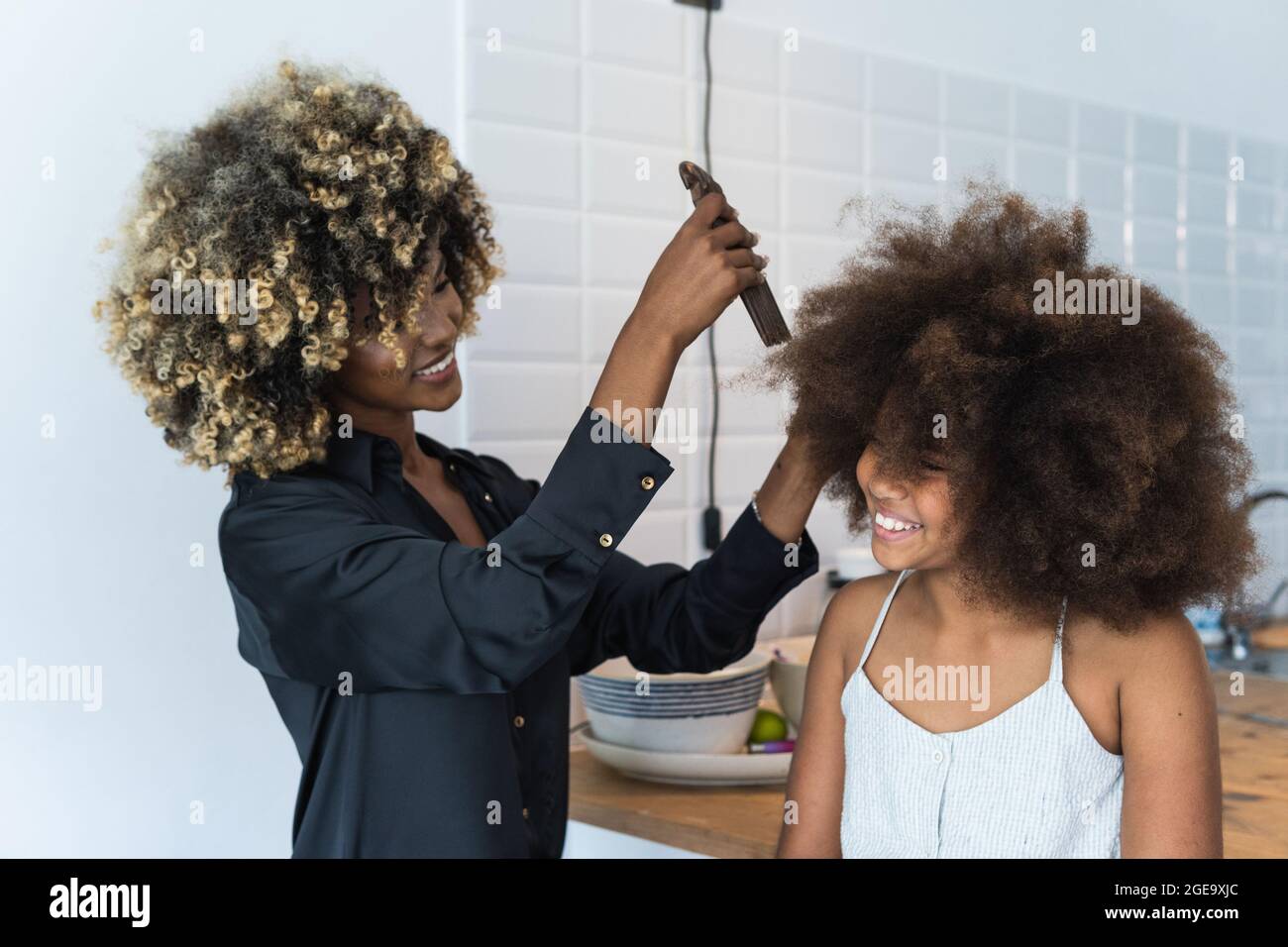 African American female using comb to do Afro hairstyle to cheerful black  girl in kitchen at home Stock Photo - Alamy