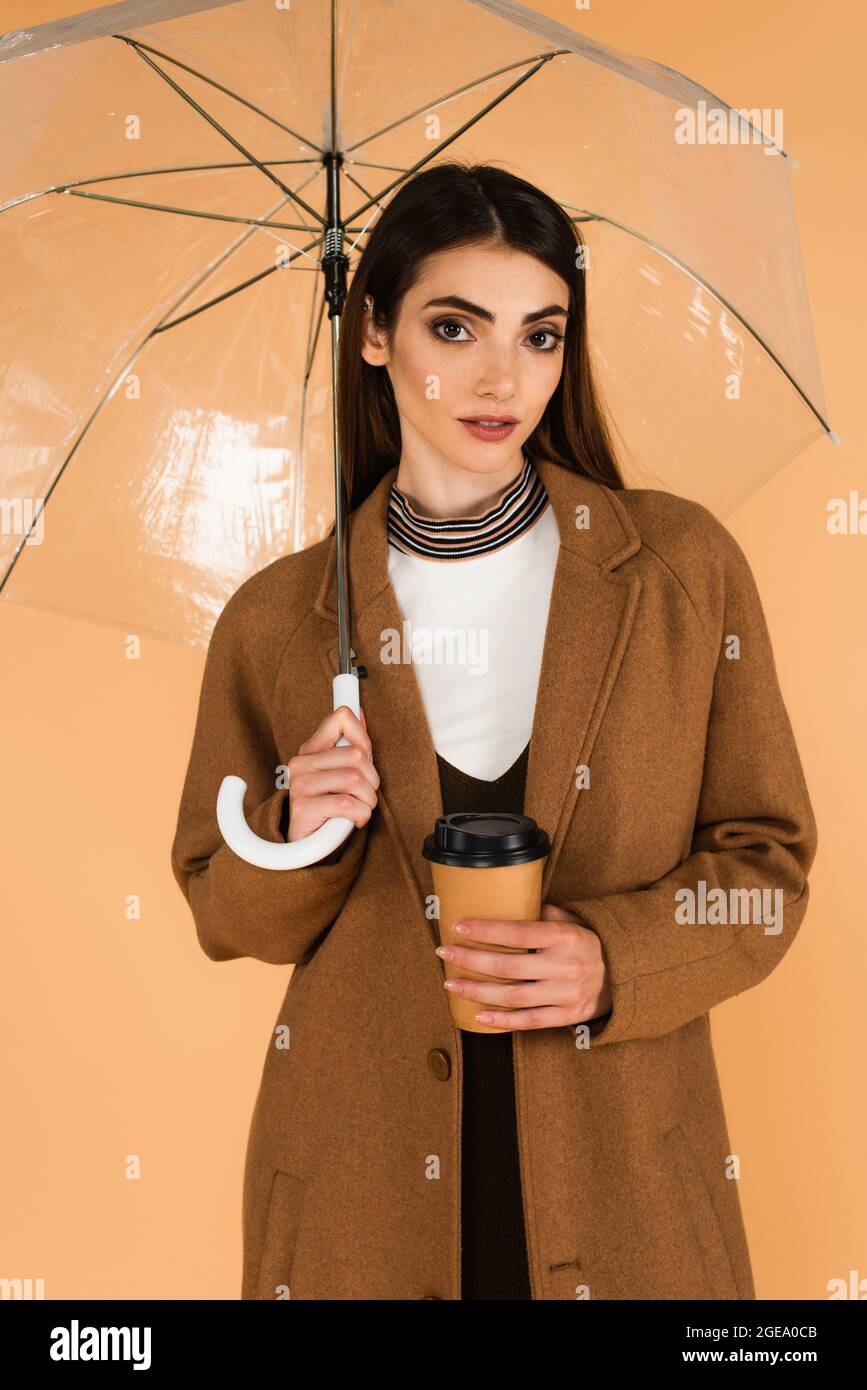 young woman in brown coat standing with paper cup under transparent umbrella isolated on beige Stock Photo