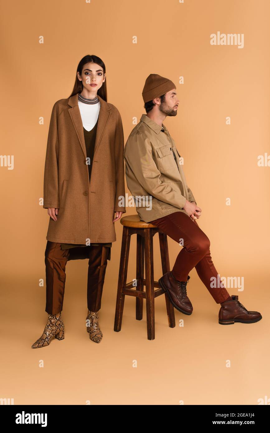 young woman in trendy coat near man sitting on stool in jacket and beanie on beige Stock Photo