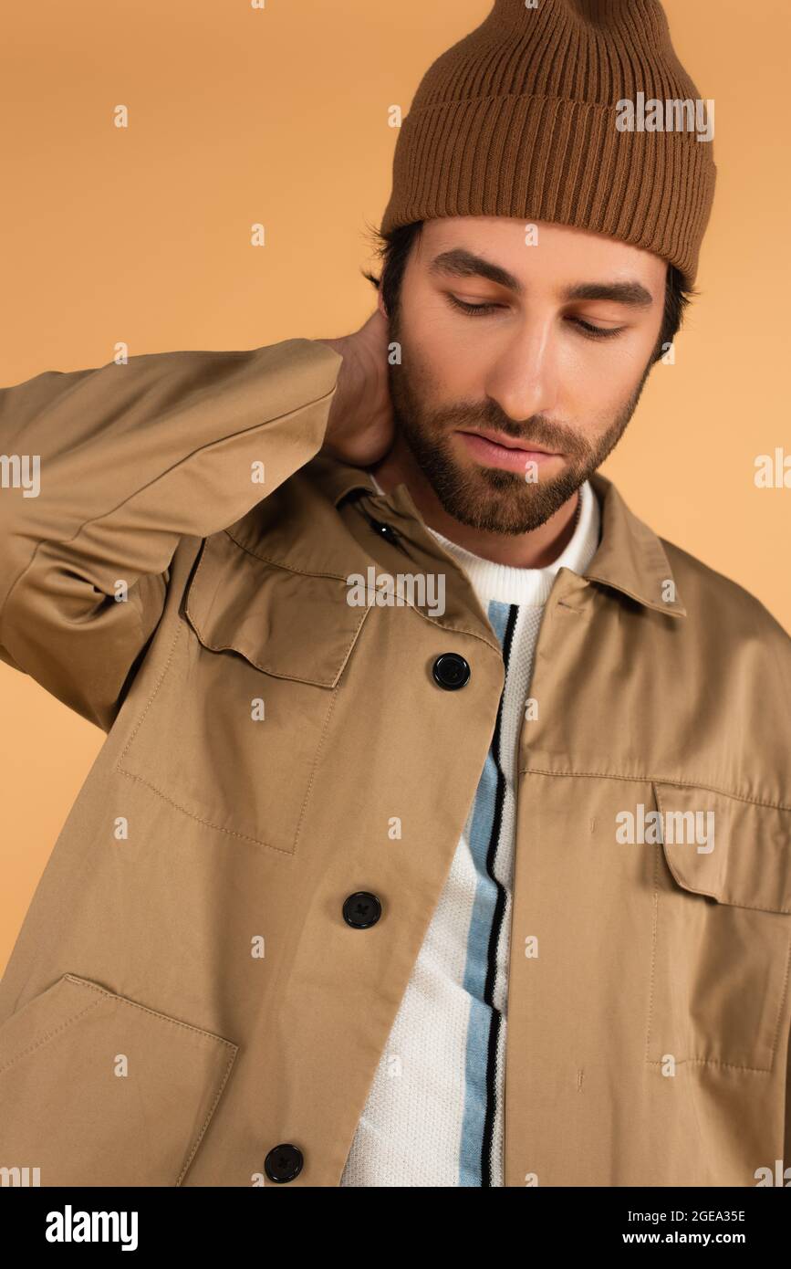 trendy man in brown beanie and jacket posing with hand on neck isolated on beige Stock Photo