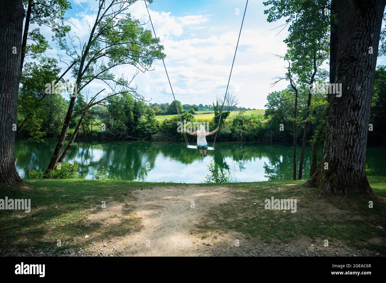 Blonde woman on a huge swing facing a green river during summer vacation - a happy day off Stock Photo