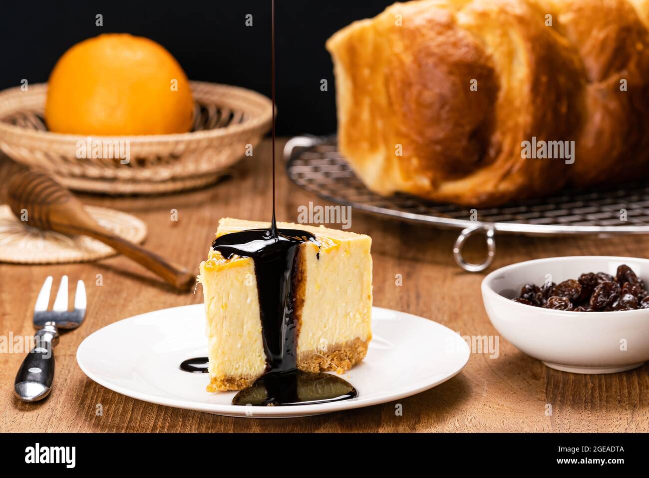 Pouring chocolate on delicious homemade mango cheesecake in white ceramic dish with metal fork, honey dipper, loaf of bread on cooling rack, ripe oran Stock Photo