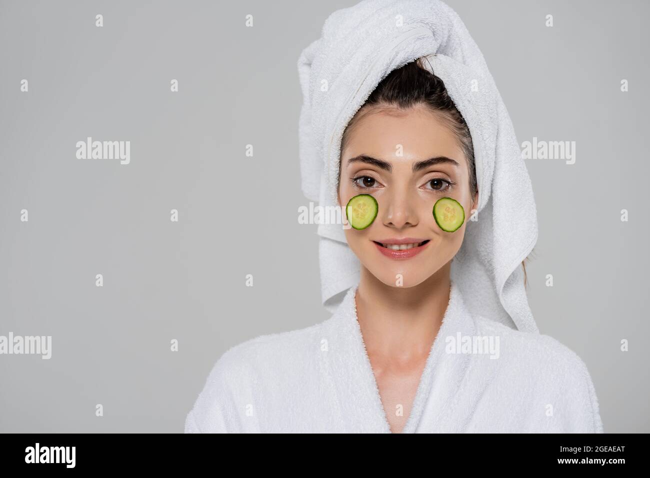 smiling young woman with towel on head and sliced cucumber on face isolated on grey Stock Photo