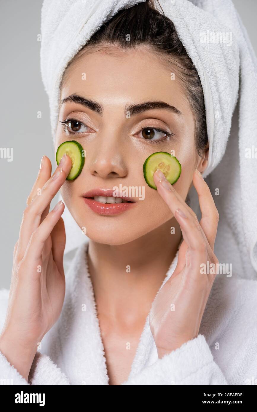 young woman with towel on head applying sliced cucumber on face isolated on grey Stock Photo