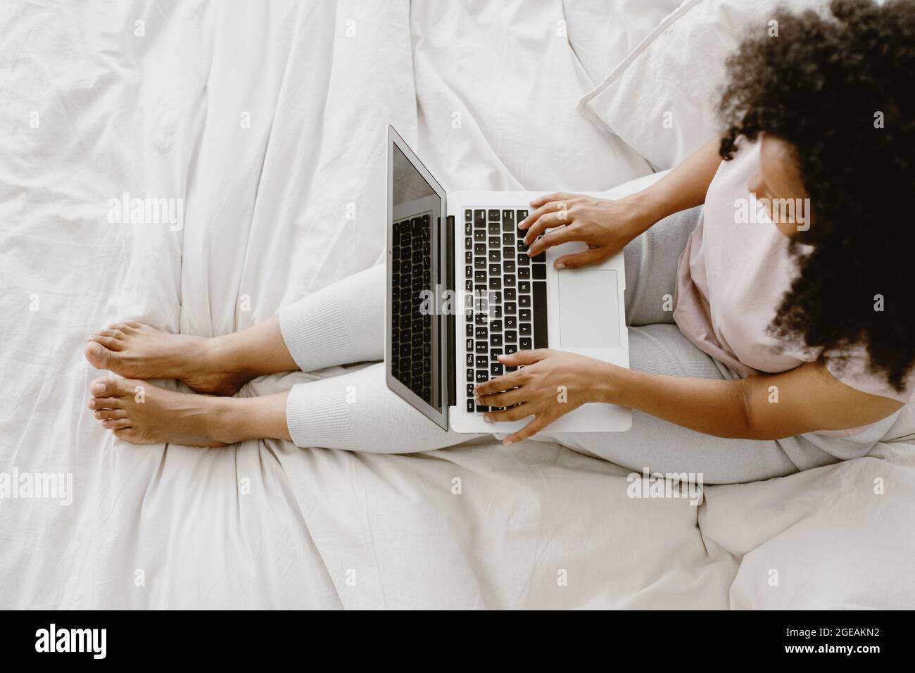 Young adorable African-American female sitting on the bed and typing on laptop. Stock Photo