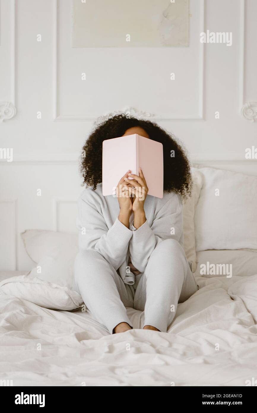 African-American woman sitting on the bed and covering face with book. Stock Photo