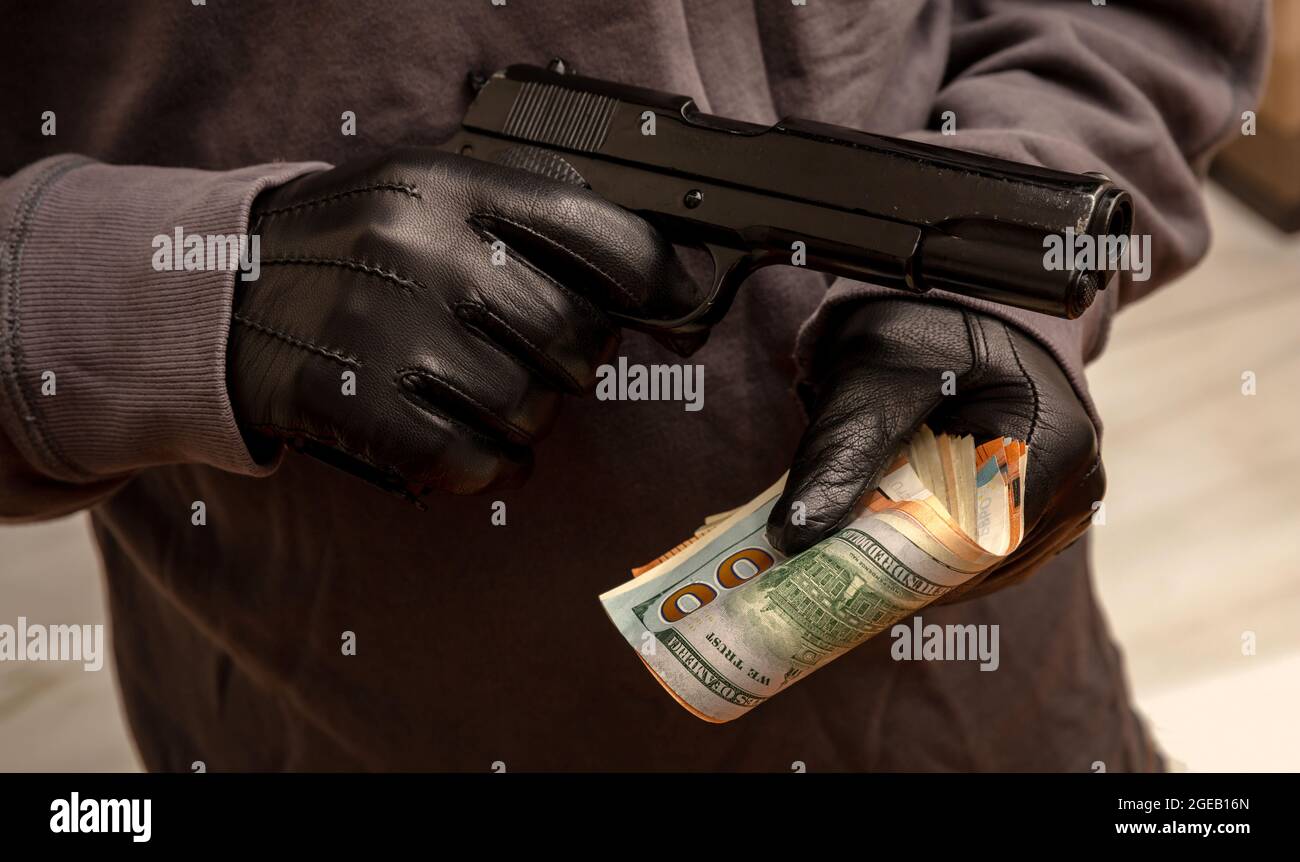 Thief threatens with a pistol for stealing money, closeup view. Armed robbery, man holding a gun and banknotes in gloved hands, Stock Photo
