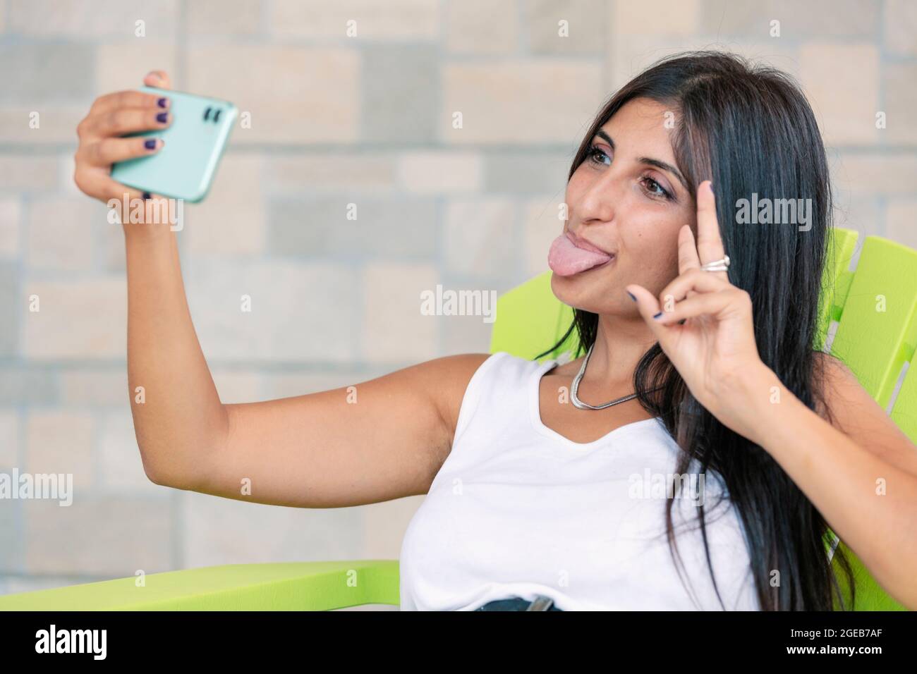 Young woman making silly face and showing tongue to smartphone while making selfie. Happy person having fun and making v sign with hand. Girl taking f Stock Photo
