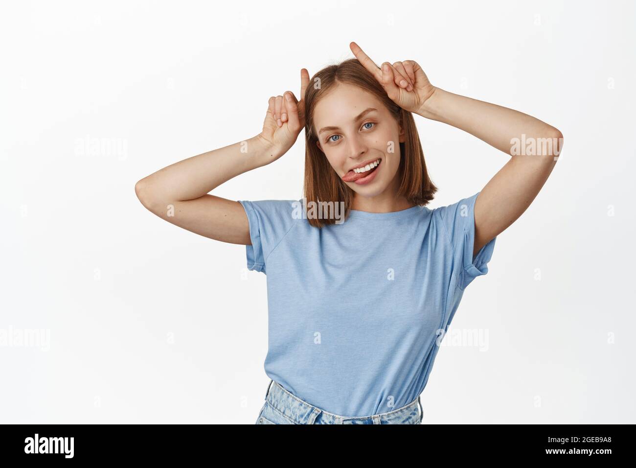 Young carefree blond woman show tongue, bull devil horns gesture on head, smiling carefree, having fun, being playful and silly, standing in t-shirt Stock Photo