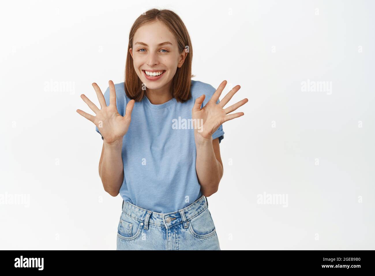 Portrait of cheerful blond girl shaking jazz hands, show empty arms and smiling excited, standing in blue t-shirt and jeans against white background Stock Photo