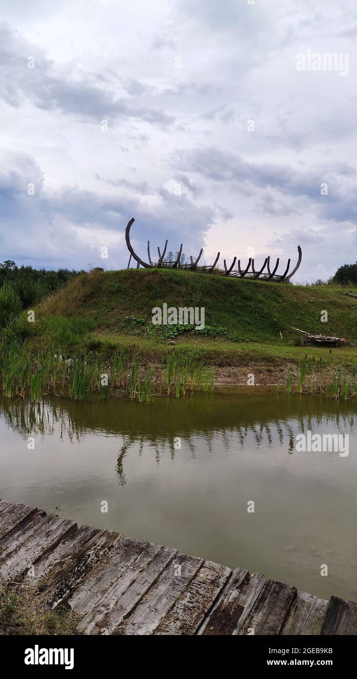 Wooden ship skeleton at the green hill near calm lake. Stock Photo