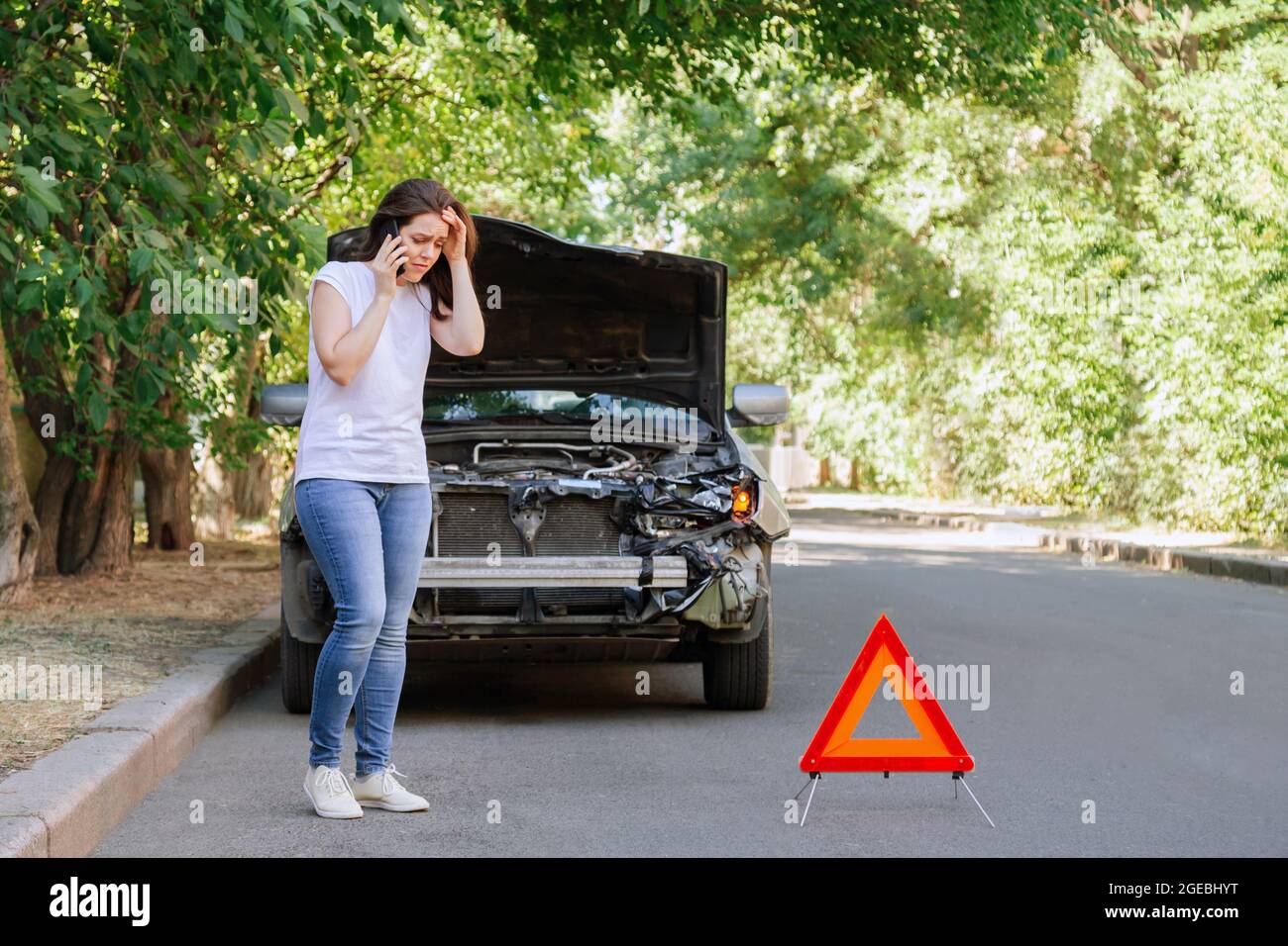 Young Woman In Front Of Broken Car In Car Accident. Woman phone calling for help and insurance after Car accident on the road with Traffic triangle Stock Photo