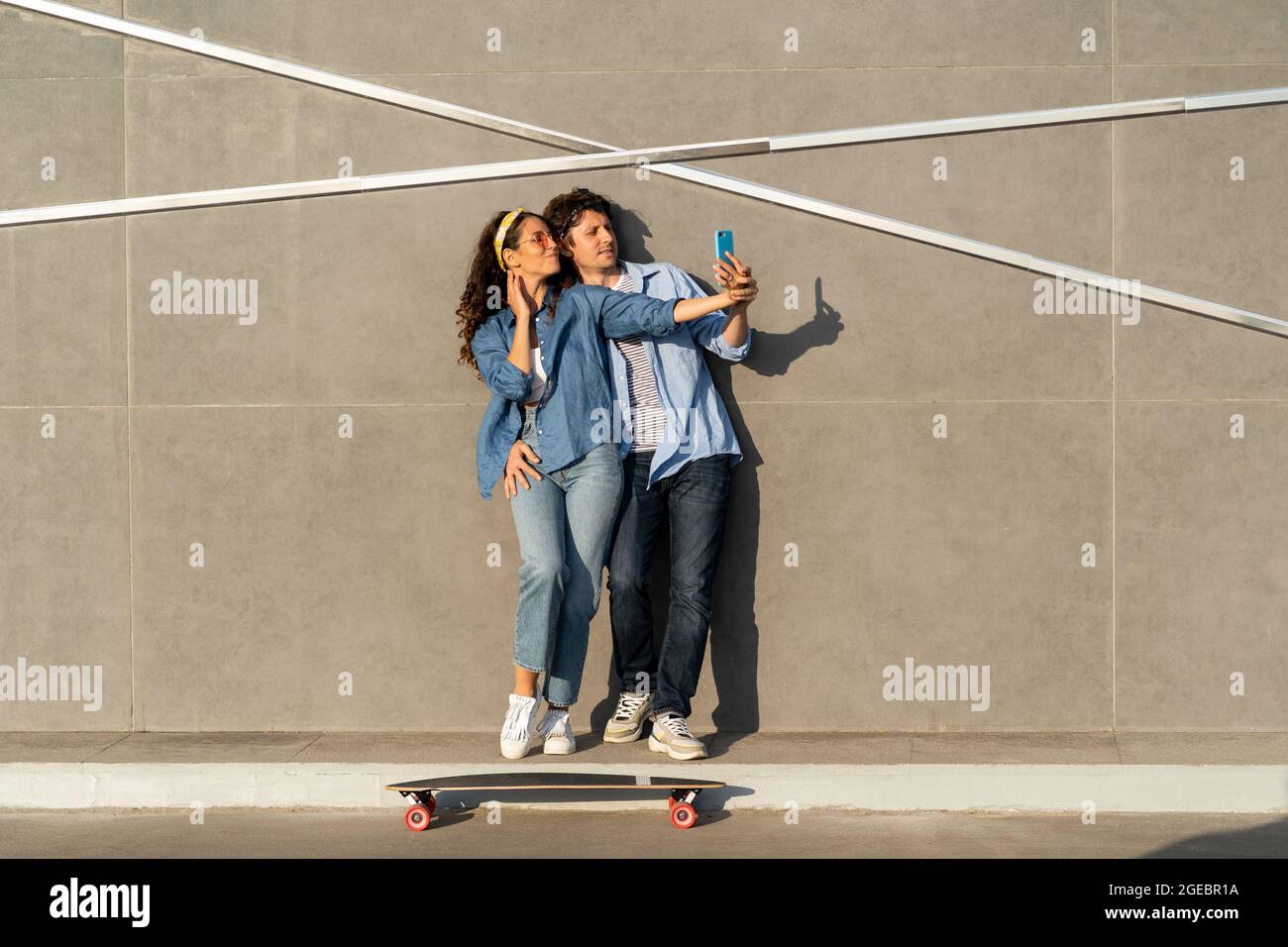 Young couple take selfie photo on smartphone over urban wall with longboard on ground happy smiling Stock Photo