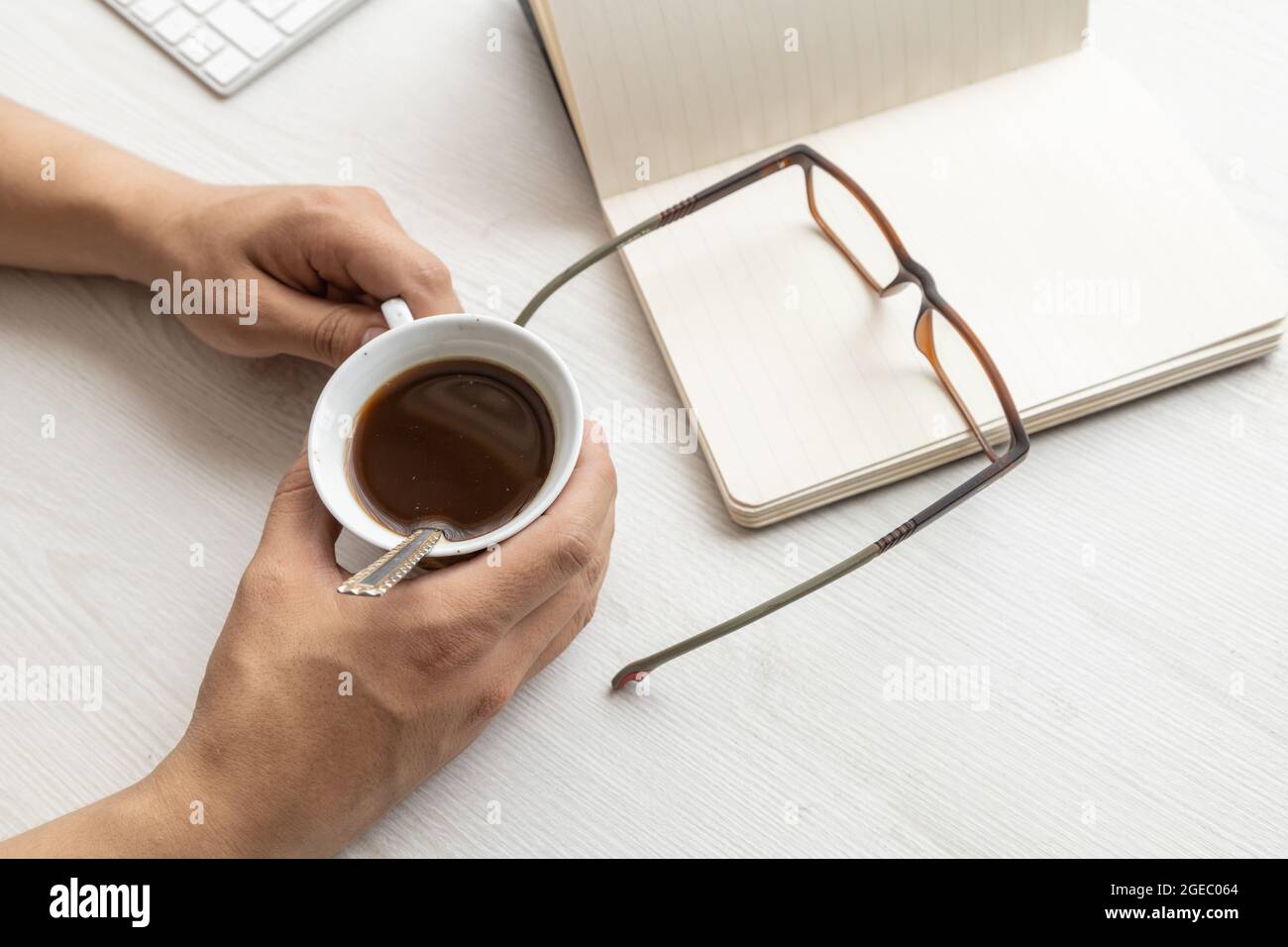 on a desk a person's hands holding a cup of coffee, with a notebook with open blank sheets and modern glasses, lifestyle in the workplace, studio Stock Photo