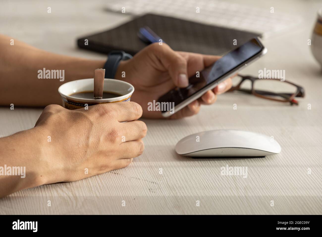 person working at his desk, detail of his hands holding a cell phone with a touch screen and with the other hand he has a cup of coffee Stock Photo