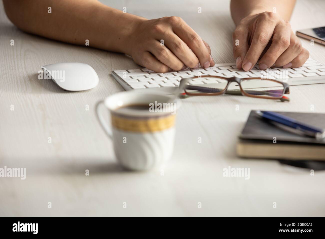 studio with a person working, detail of a person's hands typing on his modern keyboard, desk with glasses, pen, cup of coffee, computer mouse,keyboard Stock Photo
