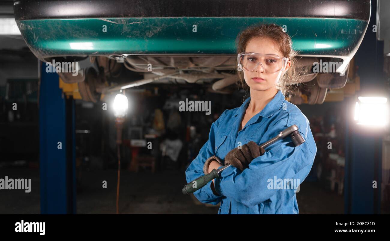 Portrait of a young beautiful Caucasian woman mechanic she stands at a car service station dressed in safety glasses and overalls. Stock Photo